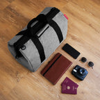 Lieblingstasche Allrounder M grey travel bag with black handles, shown with travel accessories on a wooden floor.