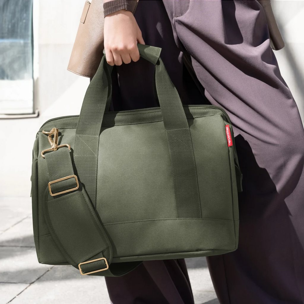Lieblingstasche Allrounder M travel bag in olive green with gold hardware, held by a person in dark trousers.