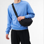 A man wearing a black Lieblingstasche sling bag across his chest with a blue sweatshirt and black trousers.