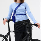 Woman wearing a bright blue Lieblingstasche sling bag across her chest while leaning on a black bicycle