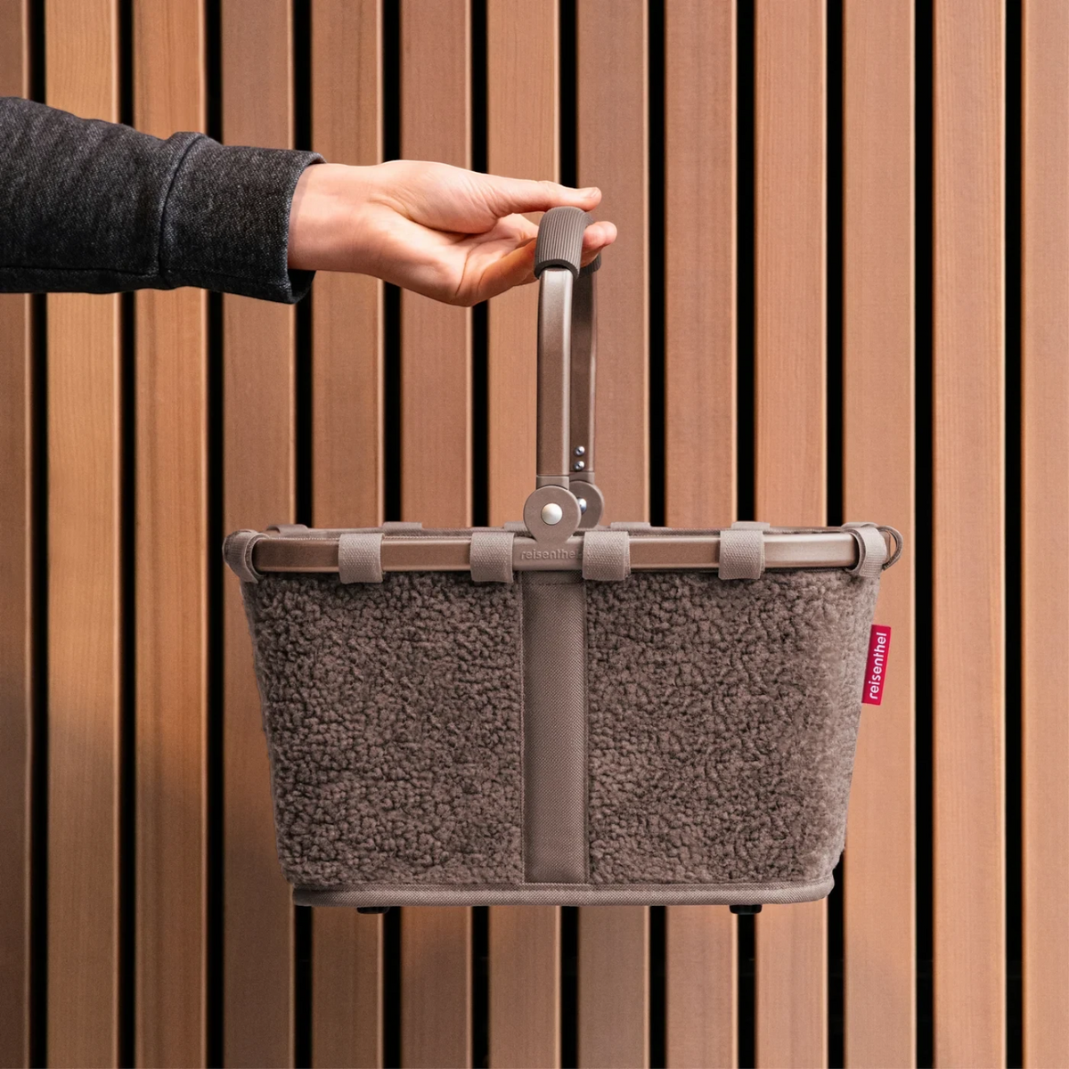 Lieblingstasche Carrybag XS in taupe teddy fleece material with a sturdy handle held against a wooden slat wall