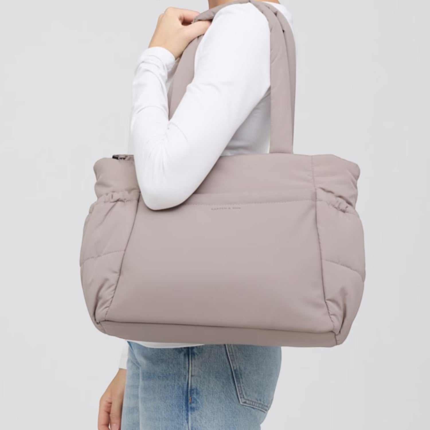 Woman carrying a taupe Lieblingstasche Hellvi Cloud Medium quilted shoulder bag against a light grey background