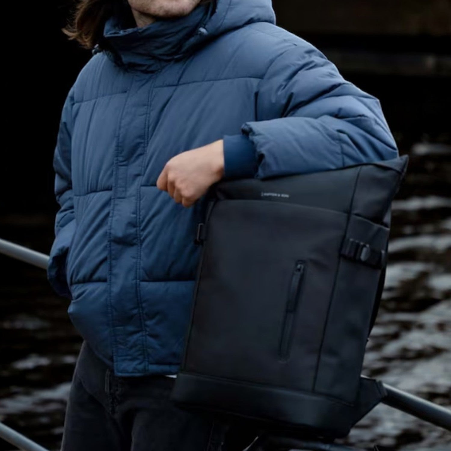 Man wearing a blue puffer jacket and black beanie posing with a Lieblingstasche Helsinki black minimalist backpack