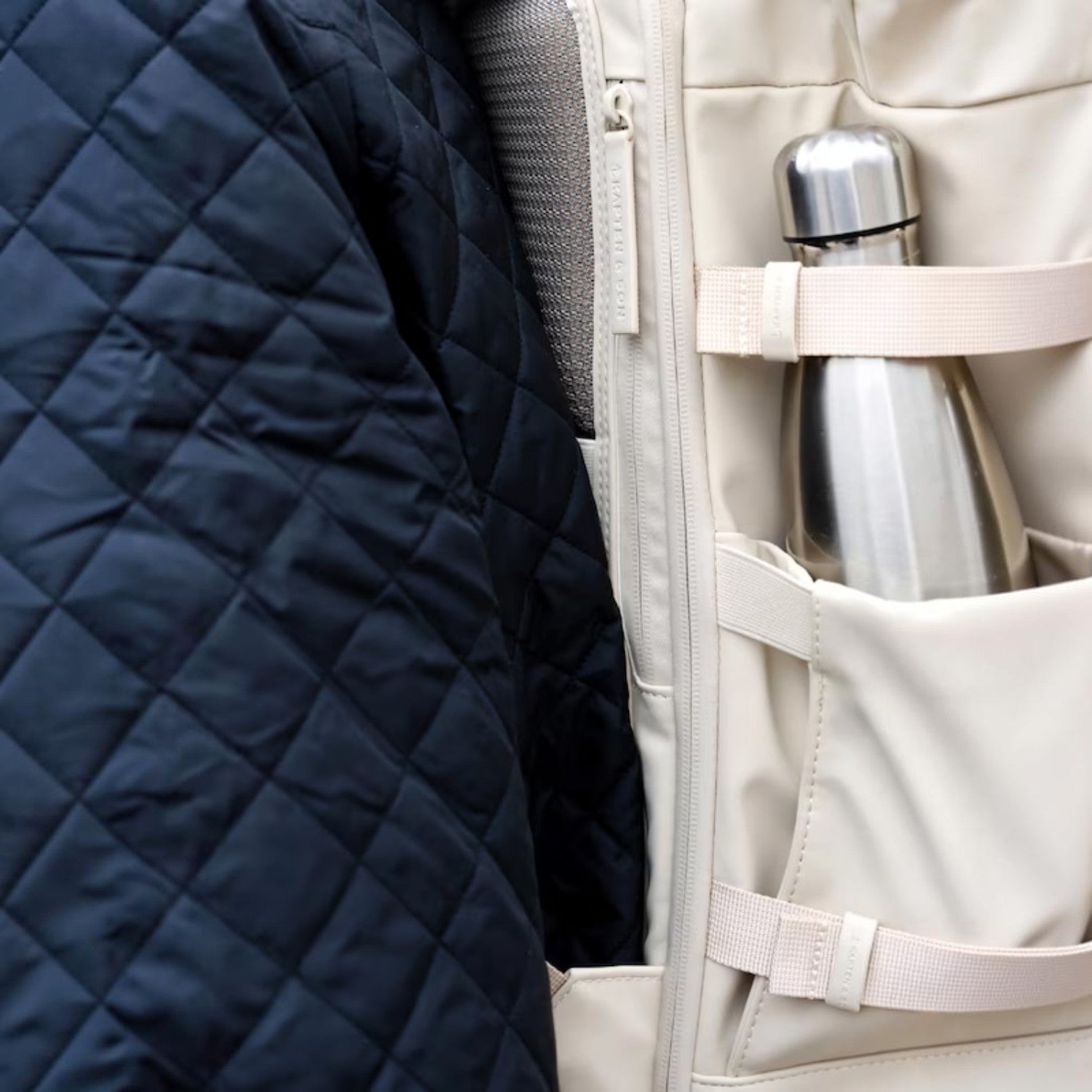 Lieblingstasche Helsinki Pro backpack in beige with a side pocket holding a silver water bottle next to a jacket