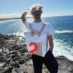 Lieblingstasche Kaje white canvas backpack with tan accents and a large orange number 2 graphic, worn by a woman overlooking the ocean.