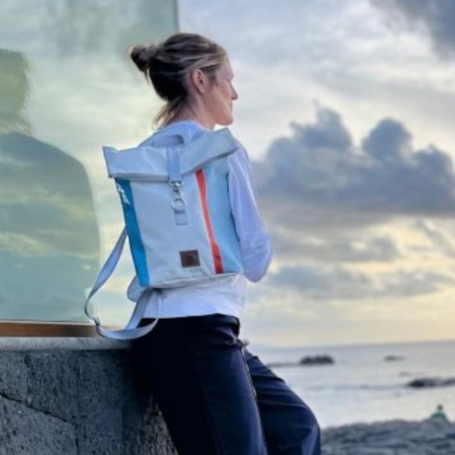 Lieblingstasche Möwe backpack in light blue and white with rolltop, worn by a woman overlooking a rocky coastline.