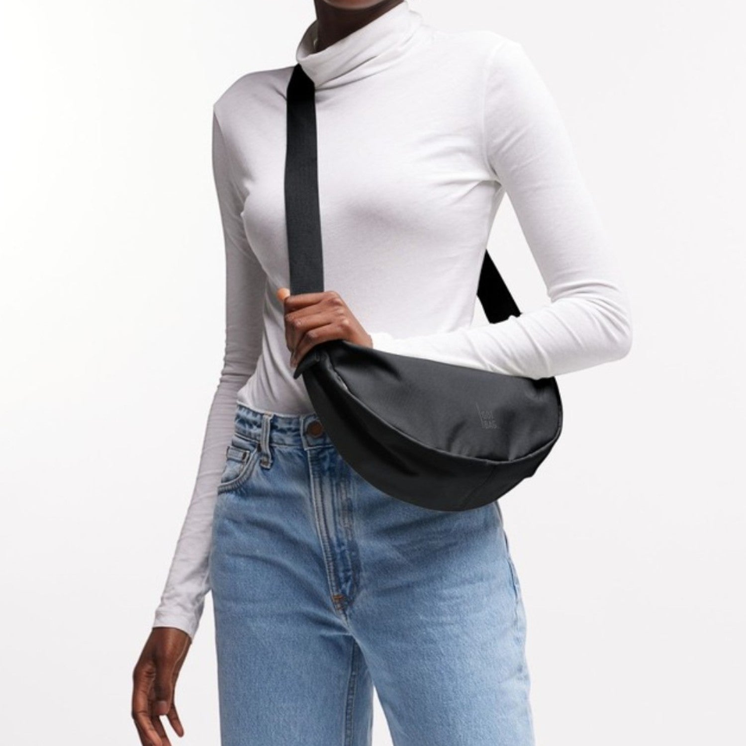 Lieblingstasche small black nylon moon bag with adjustable strap worn crossbody by a woman in a white turtleneck and jeans.