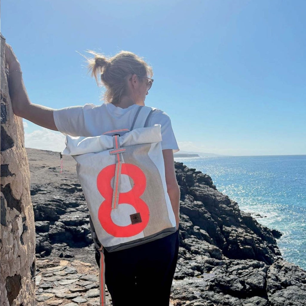 Lieblingstasche Tide roll-top backpack in white and grey with a large neon orange number eight on a coastal cliff