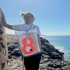Lieblingstasche Tide roll-top backpack in white and grey with a large neon orange number eight on a coastal cliff