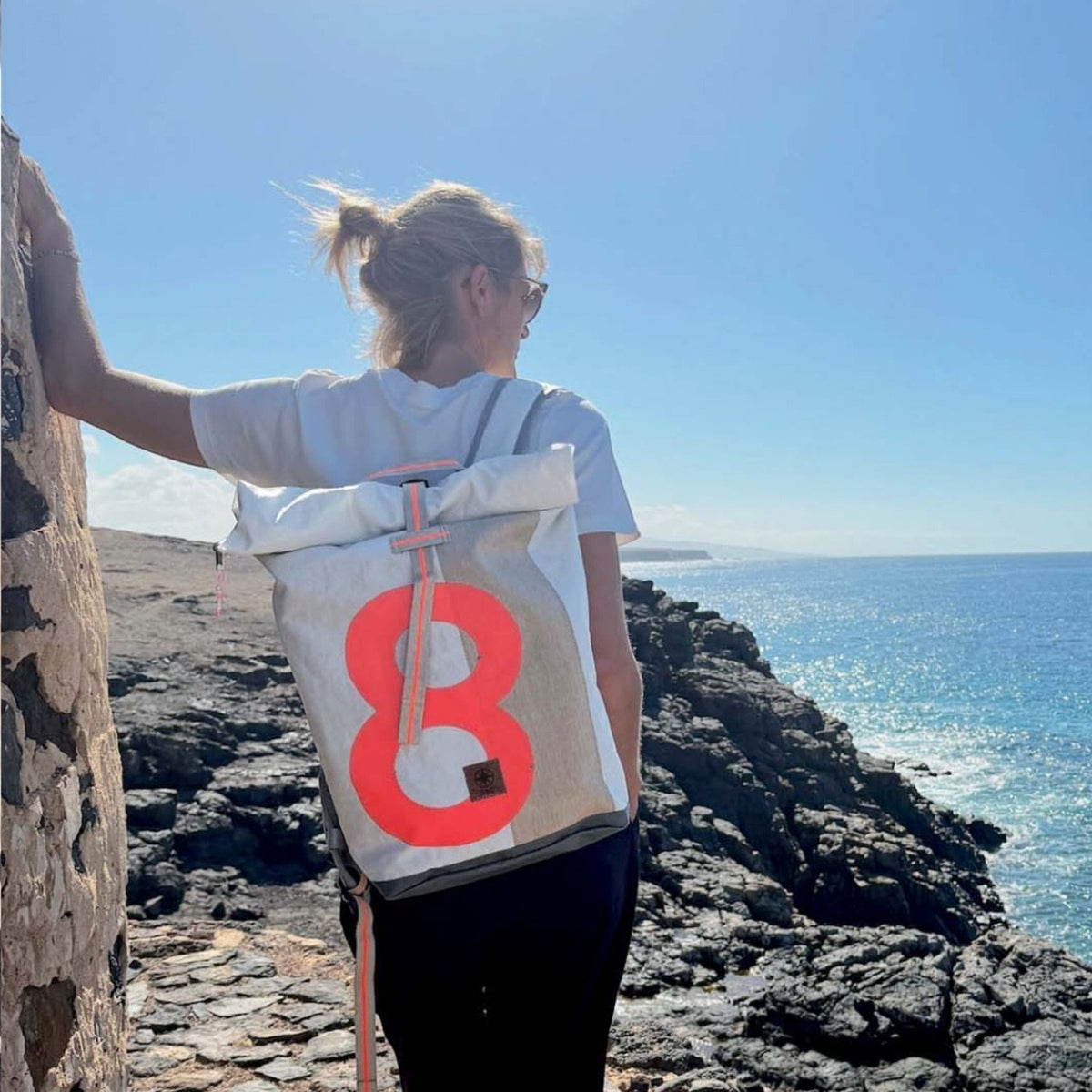 Lieblingstasche Tide roll-top backpack in white and grey with a large neon orange number eight on a coastal cliff